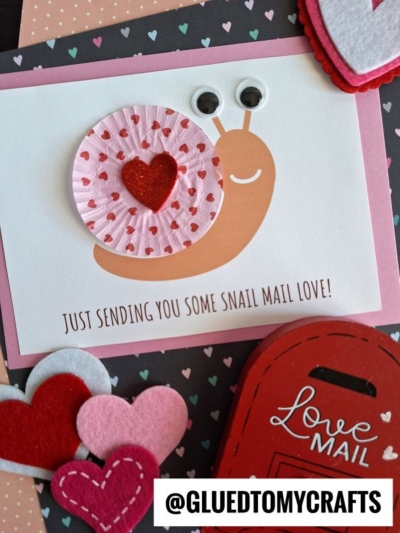 A handmade snail mail card featuring a cute cupcake liner snail with googly eyes, heart decorations, and the message “Just sending you some snail mail love!” Felt hearts and envelopes add a charming Valentine’s touch.