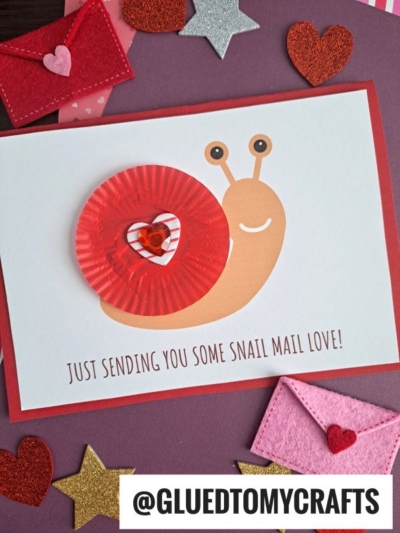 A handmade snail mail card features a smiling cupcake liner snail with a red shell, decorated with a heart. The card reads “Just sending you some SNAIL MAIL love!” and is surrounded by charming heart and star embellishments.