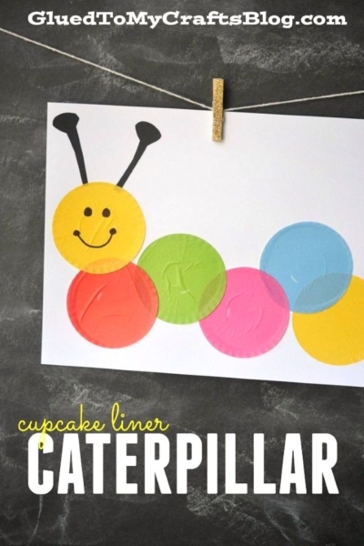 A cupcake liner caterpillar is crafted by gluing colorful cupcake liners to white paper, forming a cheerful creature with a smiling yellow face, black antennae, and a string with a clothespin at the top. CATERPILLAR is written in bold letters below.