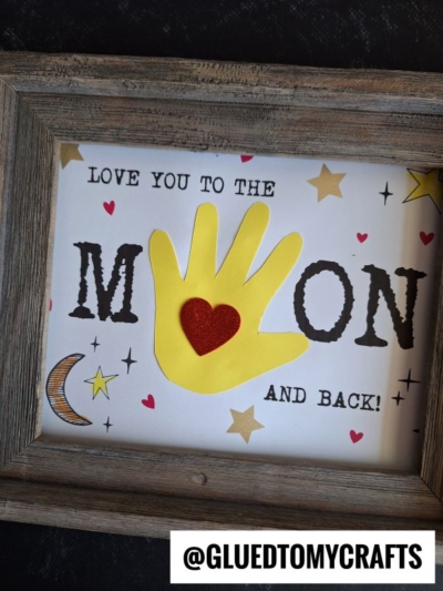 A framed craft features the phrase “handprint love you to the moon,” with a yellow paper handprint forming the O in MOON and a red heart on the palm, surrounded by stars and a crescent moon.
