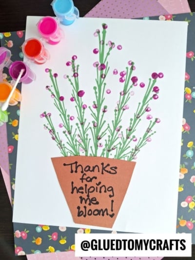 A handmade card features a flowerpot with green stems and q-tip painted lavender, created with paint and cotton swabs. The pot reads, Thanks for helping me bloom! Paint pots and paper are nearby.