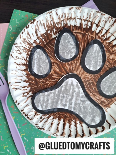 A paper plate paw print, painted with brown streaks and decorated with a large black and white design, rests on colorful confetti-patterned paper. A purple fork painted paw print sits beside it. The text @GLUEDTOMYCRAFTS appears at the bottom.
