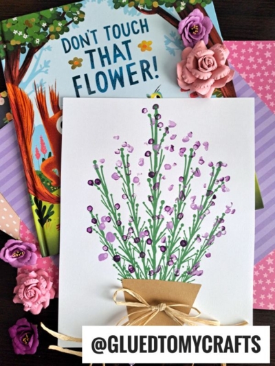 A handmade card shows q-tip painted lavender flowers in a brown pot with a bow, surrounded by pink and purple artificial flowers and the children’s book “Don’t Touch That Flower!” on a wooden surface.