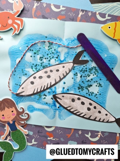 A kids fish pond craft features two paper fish attached to a popsicle stick fishing pole with string, surrounded by sea-themed stickers like a crab, fish, and mermaid, on a blue and purple patterned paint splat fishing pond background.