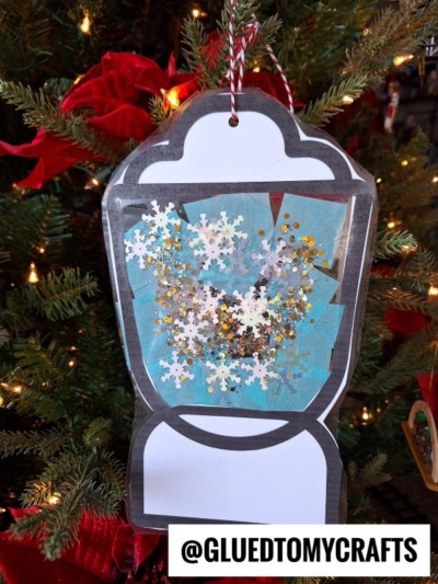 A handmade snow globe ornament with snowflakes and glitter hangs on a Christmas tree, reminiscent of the cozy glow from a stained glass camping lantern. The bottom of the ornament displays the text @GLUEDTOMYCRAFTS.