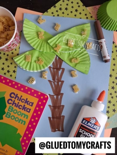 A kids craft project featuring a cupcake liner palm tree made with colorful cupcake liners and pasta letters, brown marker for the trunk, Elmer’s glue, a Chicka Chicka Boom Boom book, and a cup of letter pasta on the table.