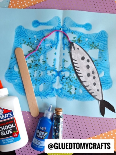 A blue fish art project is made with folded painted paper, glitter, a popsicle stick fishing pole, glue, blue paint, and yarn tail. Supplies are arranged on a pink and purple polka dot background for this fishing pond craft.