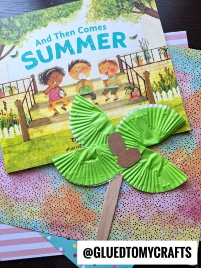 A children's book titled And Then Comes Summer is shown with a green butterfly craft made from paper cupcake liners and a wooden stick, resting on colorful, patterned paper—plus a fun cupcake liner palm tree. The handle @GLUEDTOMYCRAFTS is at the bottom.