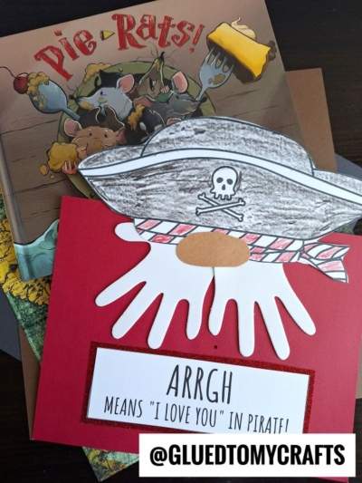 A handmade pirate handprint craft featuring a paper hat and eye patch on a white handprint, with a sign reading "ARRGH means I love you in pirate!" and the book Pie-Rats! in the background.