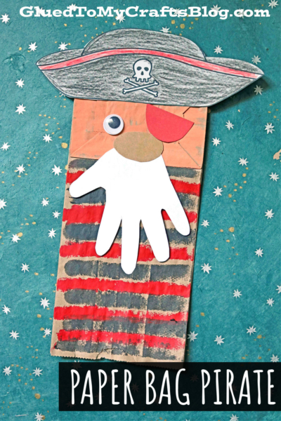 A pirate paper bag craft featuring a red and black striped shirt, a white handprint beard, a big red nose, an eye patch, and a pirate hat with skull and crossbones, all set against a star-patterned blue background.