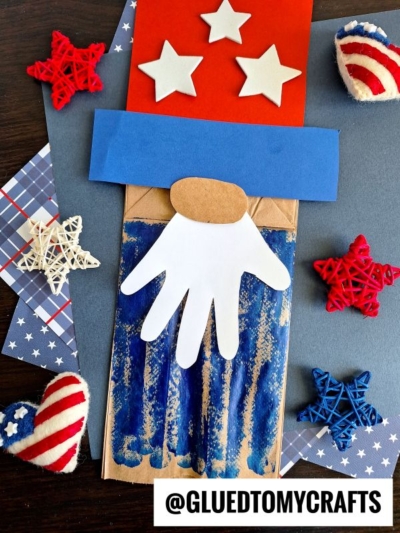 A festive paper bag Uncle Sam craft features a white handprint beard and is decorated with red, white, and blue stars and hearts. @GLUEDTOMYCRAFTS appears at the bottom.