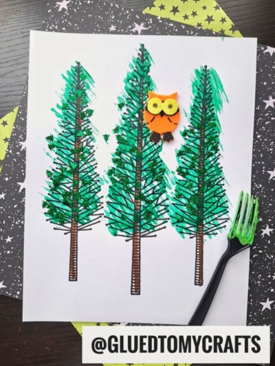 Three hand-drawn green pine trees on white paper, with a small orange owl decoration perched on the middle tree. The middle pine is a charming fork painted tree, achieved using a black plastic fork. A starry background adds to the whimsical scene. @GLUEDTOMYCRAFTS is labeled at the bottom.