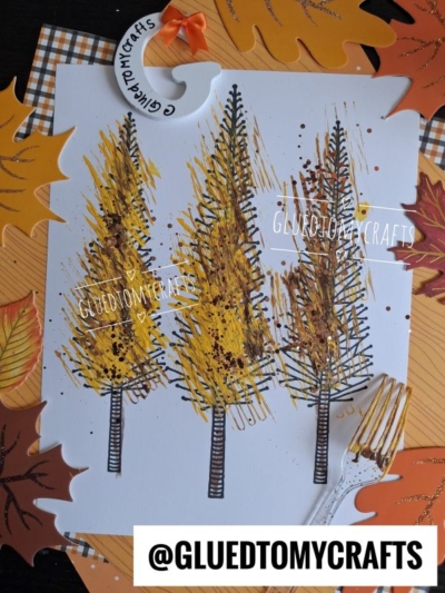Painting of three yellow autumn trees on white paper, created using the fork painted tree technique, and surrounded by artificial leaves in orange, yellow, and brown. A gold fork is placed to the right, and @GLUETOMYCRAFTS is written at the bottom.
