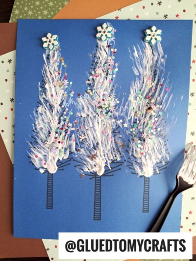 Blue paper features a trio of white fork painted pine trees, each topped with star-shaped toppers and decorated with colorful glitter ornaments. Black trunk lines add detail, while a plastic fork used for painting sits on the right. The background includes festive papers. Text: @GLUETOMYCRAFTS.