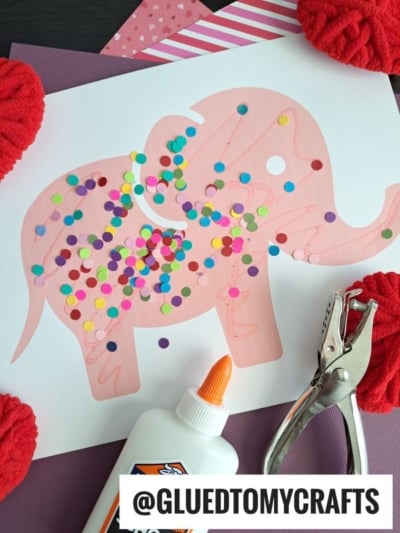 A pink paper circus animal cookie outline on white paper is decorated with colorful confetti dots. Nearby are a bottle of glue, a hole punch, red yarn, and patterned paper. The text “@GLUETOMYCRAFTS” is at the bottom.