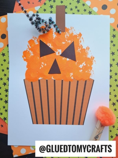 A Halloween craft features a pom pom pumpkin cupcake with orange paint, a jack-o’-lantern face, striped wrapper, and pipe cleaner stem, surrounded by star-patterned papers. A clothespin adds extra charm to the festive scene.