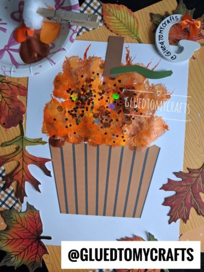 Cupcake craft made with brown striped paper, an orange sponge-painted top, colorful sequins, and a playful pom pom pumpkin cupcake accent, surrounded by fall leaves and crafting tools. A round tag and @GLUEDTOMYCRAFTS label are also visible.