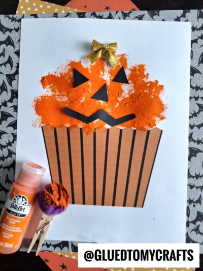A paper craft featuring a pom pom pumpkin cupcake with an orange, sponge-painted top and Jack-O’-Lantern face, black paper stripes on the base, a gold bow on top, and crafting supplies nearby. @GLUEDTOMYCRAFTS is printed on the image.