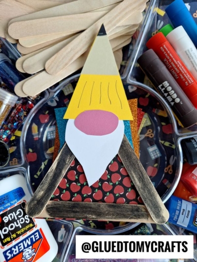 A handmade back to school gnome craft made from colored paper and popsicle sticks sits on a tray with craft supplies like popsicle sticks, glue, glitter, and markers. A label reads @GLUEDTOMYCRAFTS.