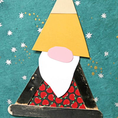 A handmade back to school gnome craft made from colored paper and popsicle sticks, featuring a yellow hat, white beard, pink nose, and red polka-dot shirt on a teal background with white stars. Text reads “Back to School Gnome Craft.”.