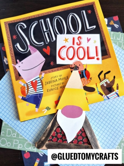 A children’s book titled School is Cool! is displayed with colorful illustrations of animals, layered over school-themed papers. In front is a handmade back to school gnome craft made of paper and craft sticks. Text reads @GLUEDTOMYCRAFTS.
