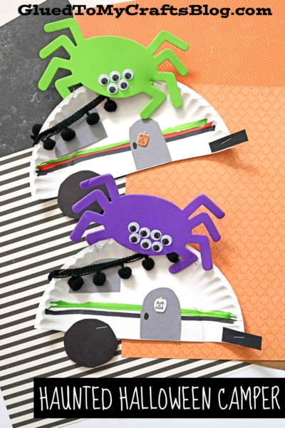 Two paper plate haunted Halloween camper crafts, decorated with green and purple foam spiders, googly eyes, black pipe cleaners, and gravestone cutouts, are displayed on patterned paper backgrounds.