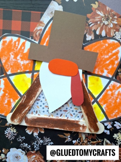 A handmade popsicle stick turkey gnome craft features a paper face with a brown hat, red and orange shapes for the wattle, decorated wings, and a triangle body. The background has charming floral and plaid patterns.
