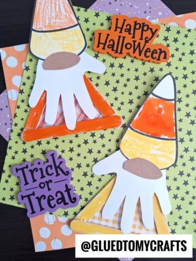 Two colorful candy corn crafts, including an adorable popsicle stick candy corn gnome made with paper handprints, are surrounded by Happy Halloween and Trick or Treat signs on orange and green star-patterned paper. Makers handle: @GLUEDTOMYCRAFTS.