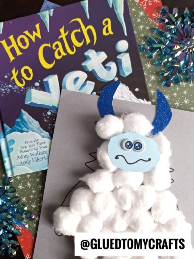A children's book titled How to Catch a Yeti sits next to a cotton ball yeti craft with blue glitter horns, placed on a decorated surface with snowflake ornaments. The handle @GLUEDTOMYCRAFTS appears at the bottom.