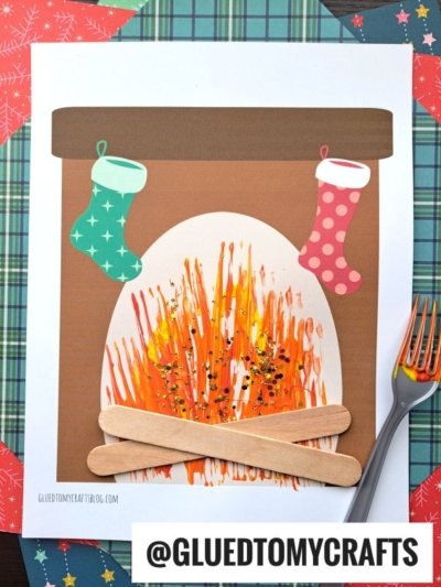 A cozy fireplace kid craft made from paper, popsicle sticks, and paint. Two stockings hang above the fork painted fireplace for a festive touch. The handle @GLUEDTOMYCRAFTS is at the bottom.