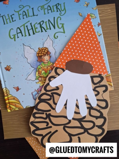 A children’s book titled “The Fall Fairy Gathering” lies on a table next to a handmade pinecone gnome with a brown acorn hat, a paper hand for a beard, and an orange polka dot hat.
