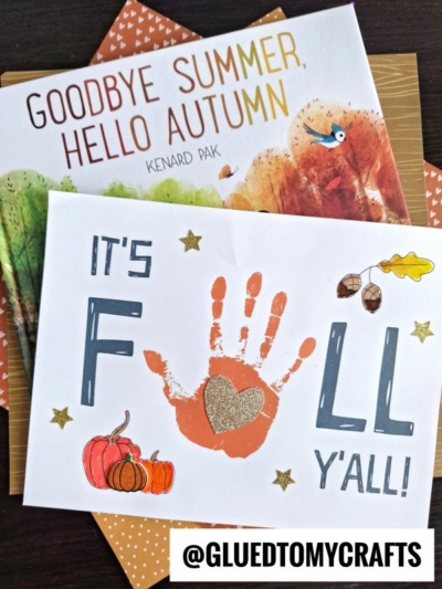 A handmade fall y'all handprint card with a child’s handprint as the “A” in “FALL,” decorated with acorns, leaves, pumpkins, and stars, sits atop the book Goodbye Summer, Hello Autumn by Kenard Pak.