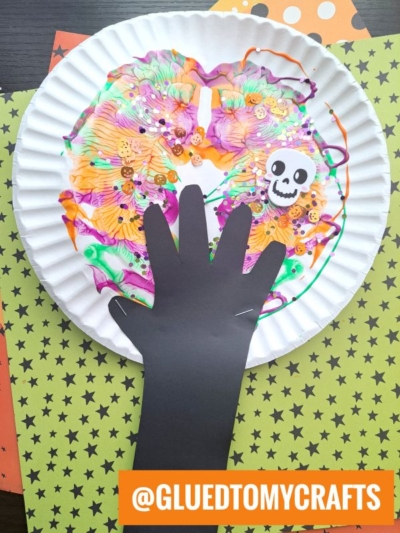 A colorful paint splat Halloween tree craft featuring a painted paper plate, black paper hand cutout, buttons, a skull sticker, and a star-patterned paper background. Text reads @GLUEDTOMYCRAFTS.