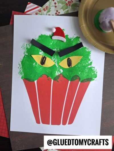 A grinch cupcake-shaped paper craft painted green to resemble the Grinch, with angry eyes, black eyebrows, and a tiny Santa hat. A green paint palette and holiday paper are nearby. Text reads @GLUEDTOMYCRAFTS. A fun grinch kid craft!.