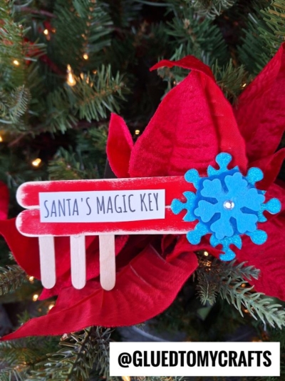 A popsicle stick Santa key labeled "Santa's Magic Key," adorned with a blue felt snowflake, hangs on a Christmas tree decorated with festive red poinsettia flowers.