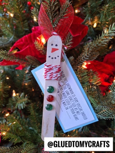 A handmade child height snowman ornament made from a craft stick with a drawn face, red and white twine scarf, and three gem buttons hangs on a Christmas tree. A gift tag and the handle @GLUEDTOMYCRAFTS are also visible.