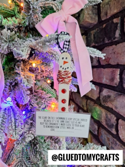 A homemade, child height snowman ornament with red buttons, a yarn scarf, and a handwritten note hangs on a snowy Christmas tree branch, decorated with a pink bow. @GLUEDTOMYCRAFTS is written at the bottom.