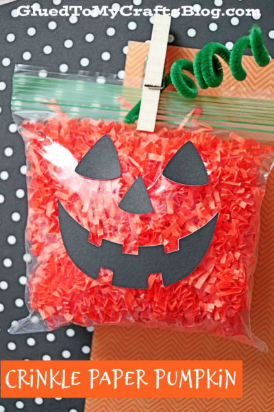 A clear ziplock bag filled with orange crinkle paper is decorated with a paper jack-o’-lantern face, green pipe cleaner stem, and labeled “Crinkle Paper Jack-O-Lantern” on an orange background.