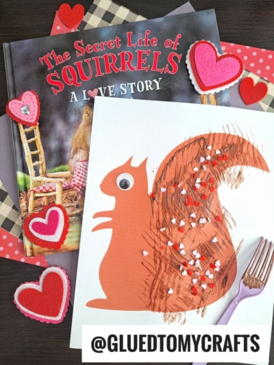 A children’s book titled The Secret Life of Squirrels: A Love Story sits beside a fork painted squirrel craft, featuring brown paint, a googly eye, and felt hearts. A @GLUEDTOMYCRAFTS label completes this charming scene.