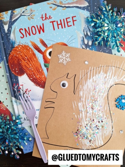A children's book titled The Snow Thief lies on a table next to a fork painted squirrel craft made from brown paper, glitter, a googly eye, a plastic fork, and blue snowflake decorations. The handle @GLUEDTOMYCRAFTS is visible.