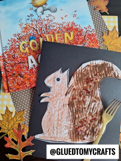 A fork painted squirrel craft with a textured tail sits atop the autumn-themed book The Golden Acorn, surrounded by faux fall leaves, a yellow plastic fork, and a gingham napkin. @GLUEDTOMYCRAFTS is written at the bottom.