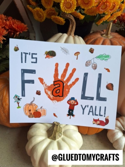 A fall-themed craft sign features “It’s FALL Y’ALL” with a bright orange handprint as the A in FALL, making it a unique handprint fall y'all project, adorned with autumn stickers and surrounded by pumpkins and flowers.
