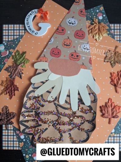 A fall-themed pinecone gnome craft made with paper, a handprint beard, pumpkin hat, and glitter, surrounded by autumn leaves and plaid paper. Text reads @GLUEDTOMYCRAFTS.