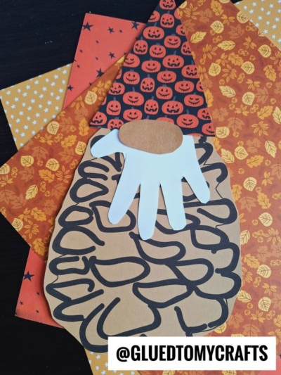 A charming paper turkey craft made from hand and oval cutouts is displayed on autumn-themed scrapbook paper with fall leaves, stars, and pumpkins. The cozy scene brings to mind festive pinecone gnome decorations, perfect for seasonal crafting. The @GLUEDTOMYCRAFTS watermark is at the bottom.
