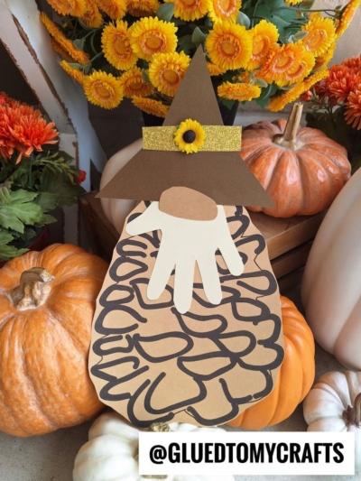 A paper gnome craft with a brown hat, sunflower accent, and handprint beard sits among orange pumpkins, vibrant fall flowers, and a charming pinecone gnome. @GLUEDTOMYCRAFTS appears at the bottom of the image.