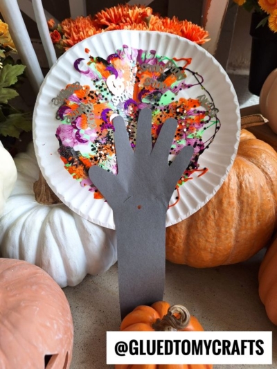 A paper plate decorated with a vibrant paint splat and glitter stands behind a black paper cut-out of a Halloween tree hand, surrounded by pumpkins. Text at the bottom reads @GLUEDTOMYCRAFTS.