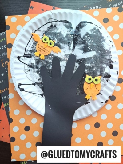 A Halloween craft featuring a black paper hand cutout as a paint splat Halloween tree on a white paper plate, with orange felt owls, set on polka dot and Halloween-themed papers. Text reads @GLUEDTOMYCRAFTS.