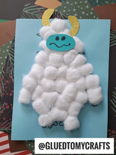 A cute cotton ball yeti craft made from fluffy cotton balls glued to blue paper, with a blue face, black smile, and gold horns. The image also features the text @GLUEDTOMYCRAFTS at the bottom.