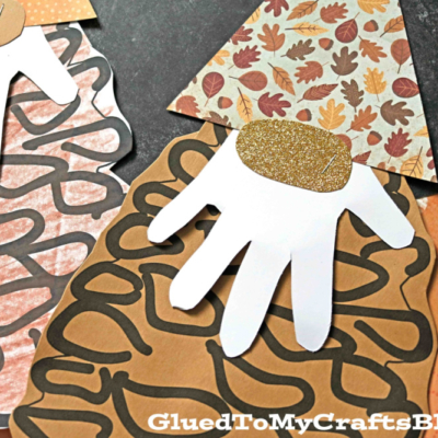 A handmade pine cone gnome crafted from cut-out paper shapes, featuring a textured pinecone body, white handprint beard, peach oval nose, and orange polka-dot hat.