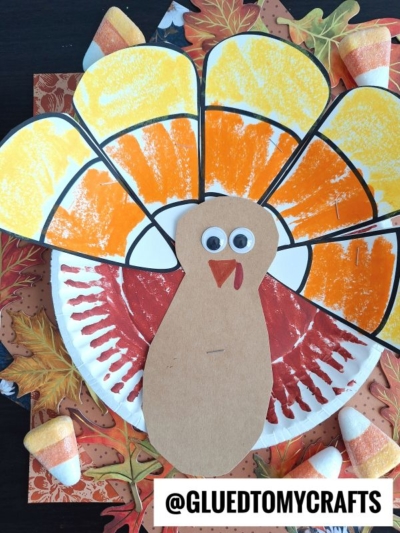 A handmade paper plate candy corn turkey craft featuring a painted paper plate body, a colorful feather fan, a brown paper head with googly eyes, and candy corn accents on a leafy surface. A watermark reads @GLUEDTOMYCRAFTS.
